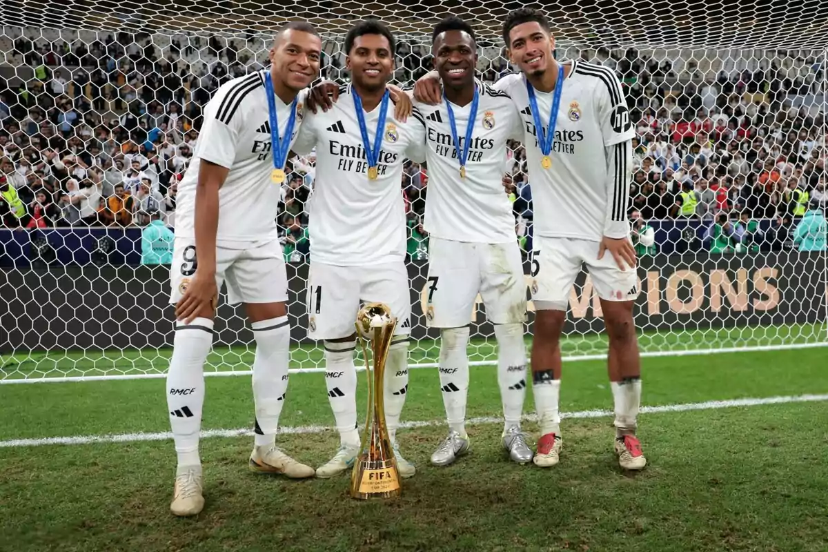 Cuatro jugadores de fútbol del Real Madrid posan juntos en el campo con medallas y un trofeo dorado frente a ellos, con una multitud de espectadores al fondo.