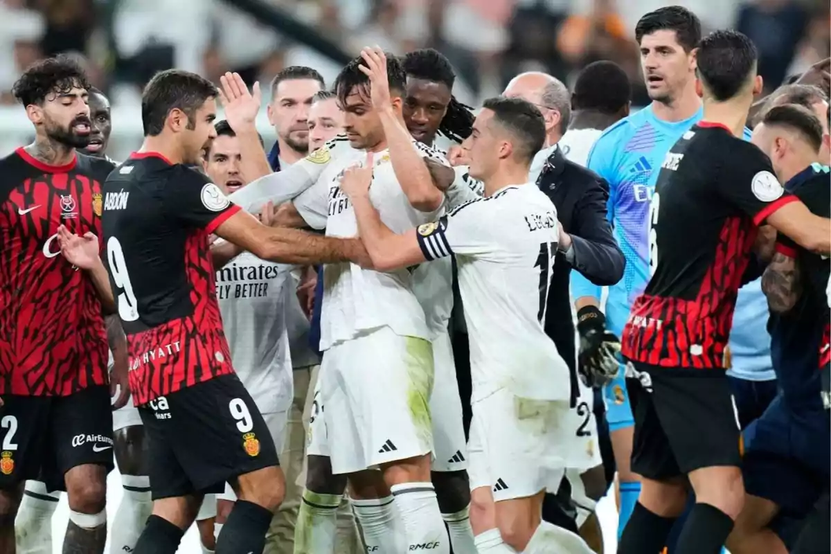 Jugadores de dos equipos de fútbol discuten en el campo rodeados de compañeros y árbitros.