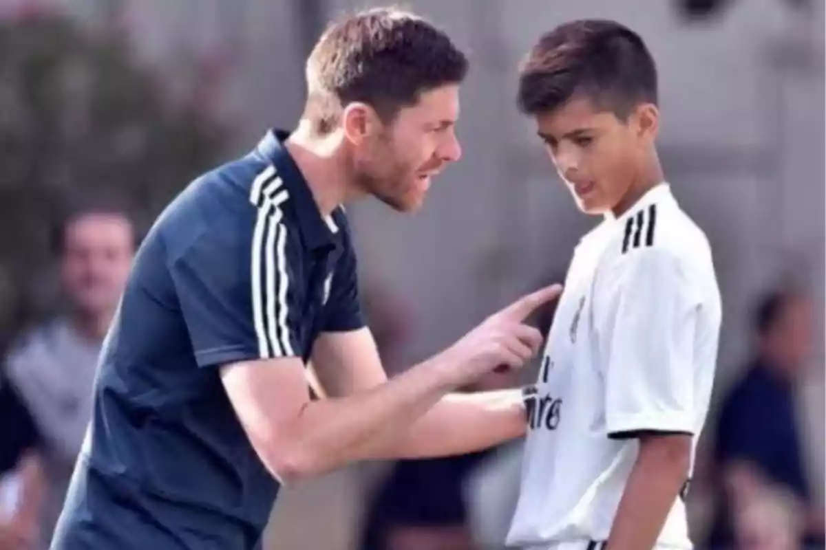 A man in a dark blue sports uniform seems to be giving instructions to a young man in a white uniform on a training field. A man in a dark blue sports uniform seems to be giving instructions to a young man in a white uniform on a training field.