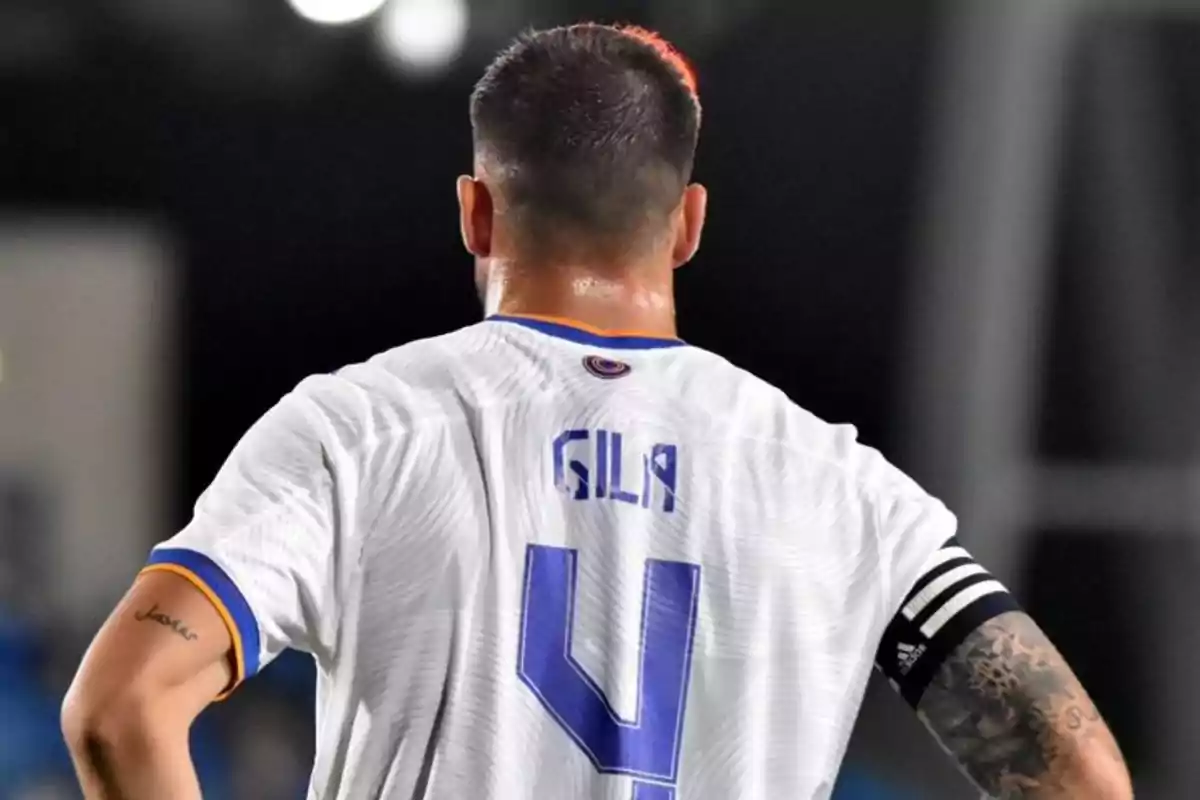 Soccer player seen from behind with the number 4 and the name Gila on the white jersey, wearing a captain's armband and showing tattoos on both arms Soccer player seen from behind with the number 4 and the name Gila on the white jersey, wearing a captain's armband and showing tattoos on both arms