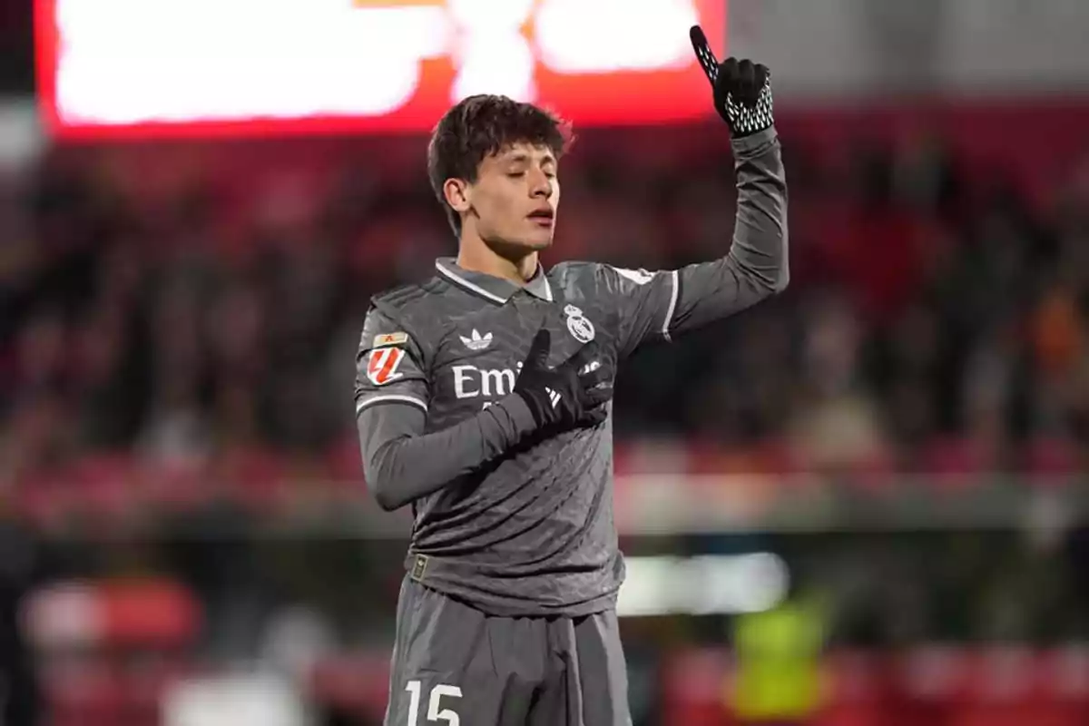 A soccer player in a gray uniform raises one arm and holds the other hand on his chest in a stadium.