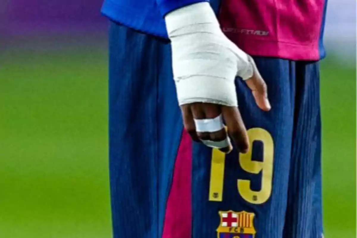 Un jugador de fútbol con la mano vendada y cinta en los dedos lleva el uniforme del FC Barcelona con el número 19. Un jugador de fútbol con la mano vendada y cinta en los dedos lleva el uniforme del FC Barcelona con el número 19.