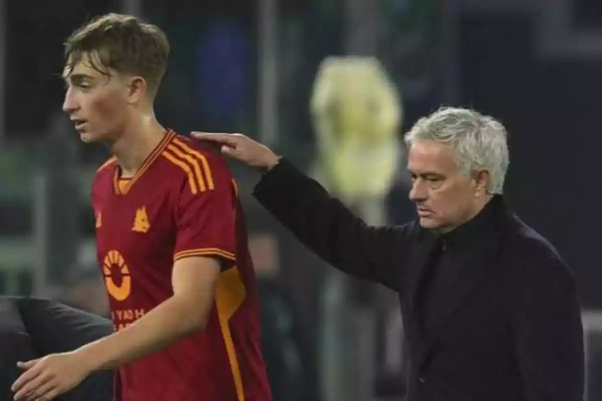 Un hombre con uniforme de fútbol rojo es tocado en el hombro por otro hombre de cabello canoso y abrigo oscuro en un entorno deportivo. Un hombre con uniforme de fútbol rojo es tocado en el hombro por otro hombre de cabello canoso y abrigo oscuro en un entorno deportivo.