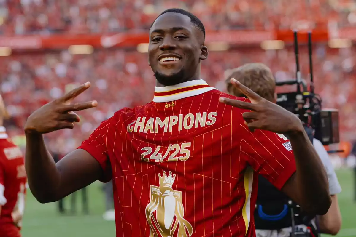 Un jugador de fútbol sonríe y hace el signo de la paz con ambas manos mientras lleva una camiseta roja con la palabra Un jugador de fútbol sonríe y hace el signo de la paz con ambas manos mientras lleva una camiseta roja con la palabra