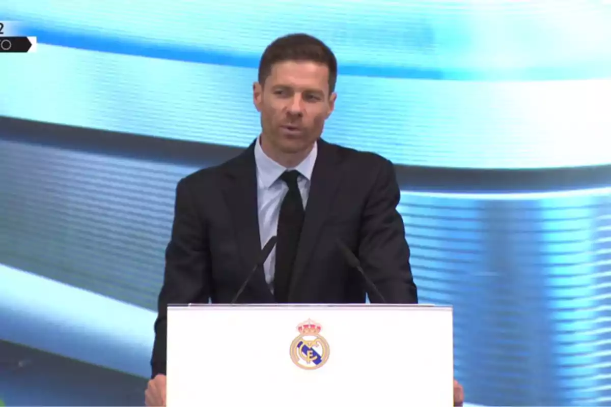 Man in a suit speaking at a podium with the Real Madrid crest on the front and a light blue background. Man in a suit speaking at a podium with the Real Madrid crest on the front and a light blue background.
