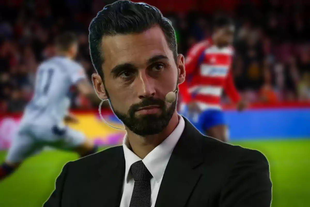 Bearded man with a headset microphone wearing a suit and tie in the foreground with a blurred soccer match in the background