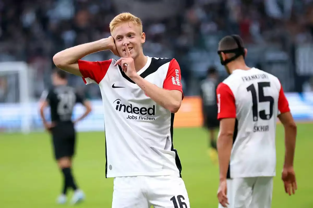 A player celebrates a goal by making a shushing gesture with his finger on his lips while another player walks behind him on the field. A player celebrates a goal by making a shushing gesture with his finger on his lips while another player walks behind him on the field.