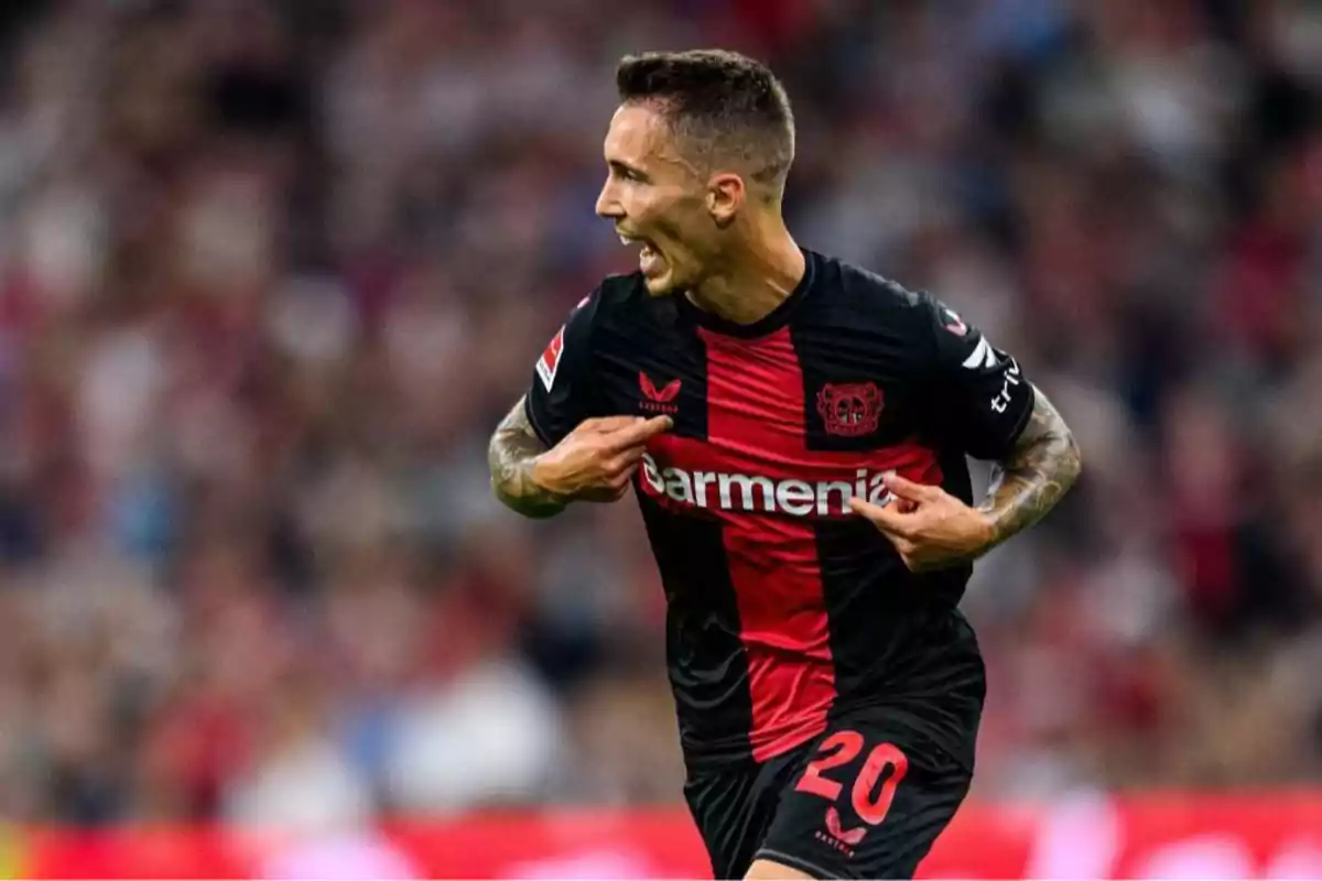 A player celebrating a goal while pointing to the badge on his Bayer Leverkusen jersey in a stadium full of spectators. A player celebrating a goal while pointing to the badge on his Bayer Leverkusen jersey in a stadium full of spectators.