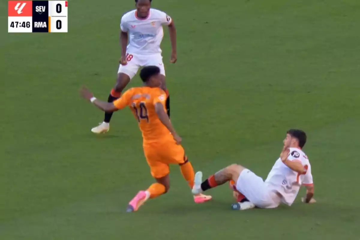 Un jugador de fútbol con uniforme naranja está en movimiento mientras otro jugador con uniforme blanco está en el suelo durante un partido. Un jugador de fútbol con uniforme naranja está en movimiento mientras otro jugador con uniforme blanco está en el suelo durante un partido.