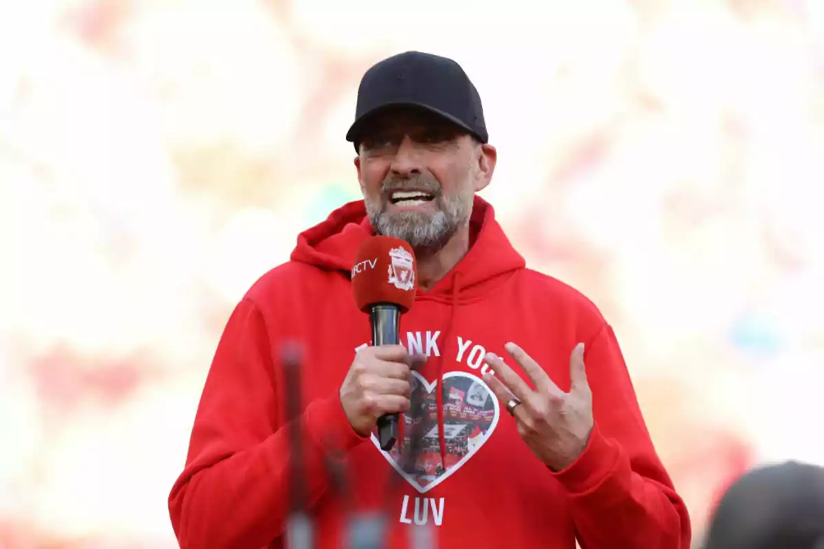 A man with a beard and a black cap holds a red microphone while wearing a red sweatjersey with a heart design.