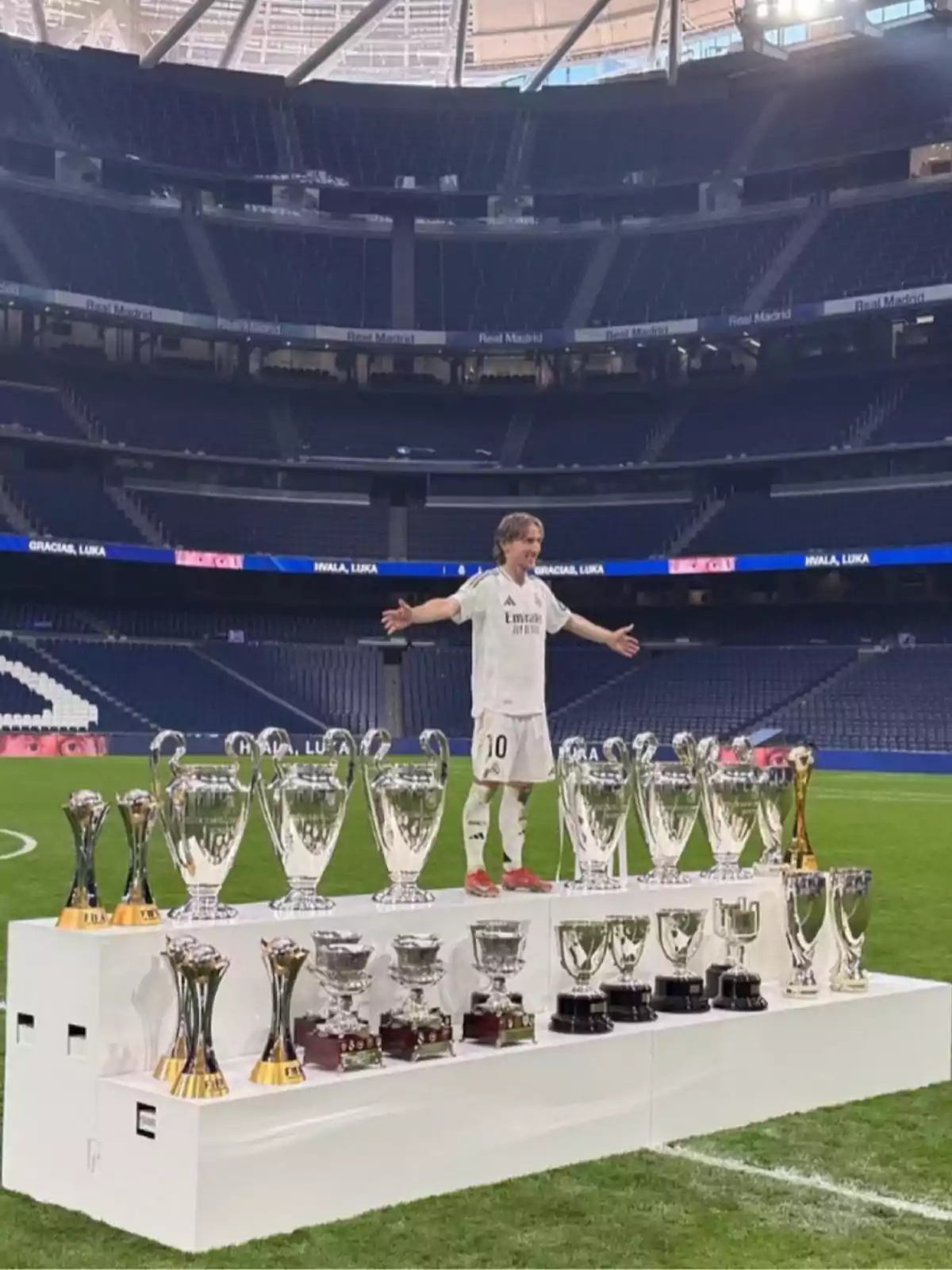Un jugador de fútbol con uniforme blanco está de pie en un estadio vacío, rodeado de numerosos trofeos en una plataforma blanca. Un jugador de fútbol con uniforme blanco está de pie en un estadio vacío, rodeado de numerosos trofeos en una plataforma blanca.