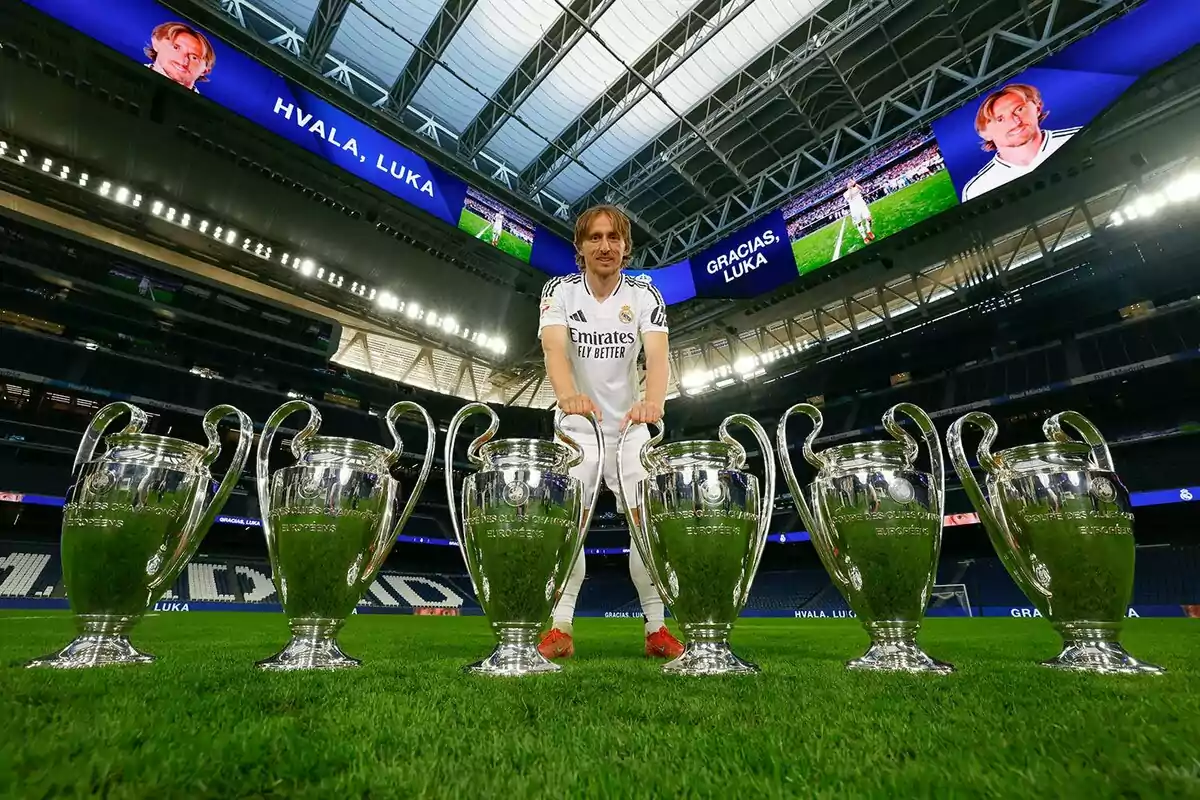 Un jugador de fútbol posa en un estadio con varios trofeos de la Liga de Campeones alineados frente a él, mientras en las pantallas del estadio se muestra un mensaje de agradecimiento.