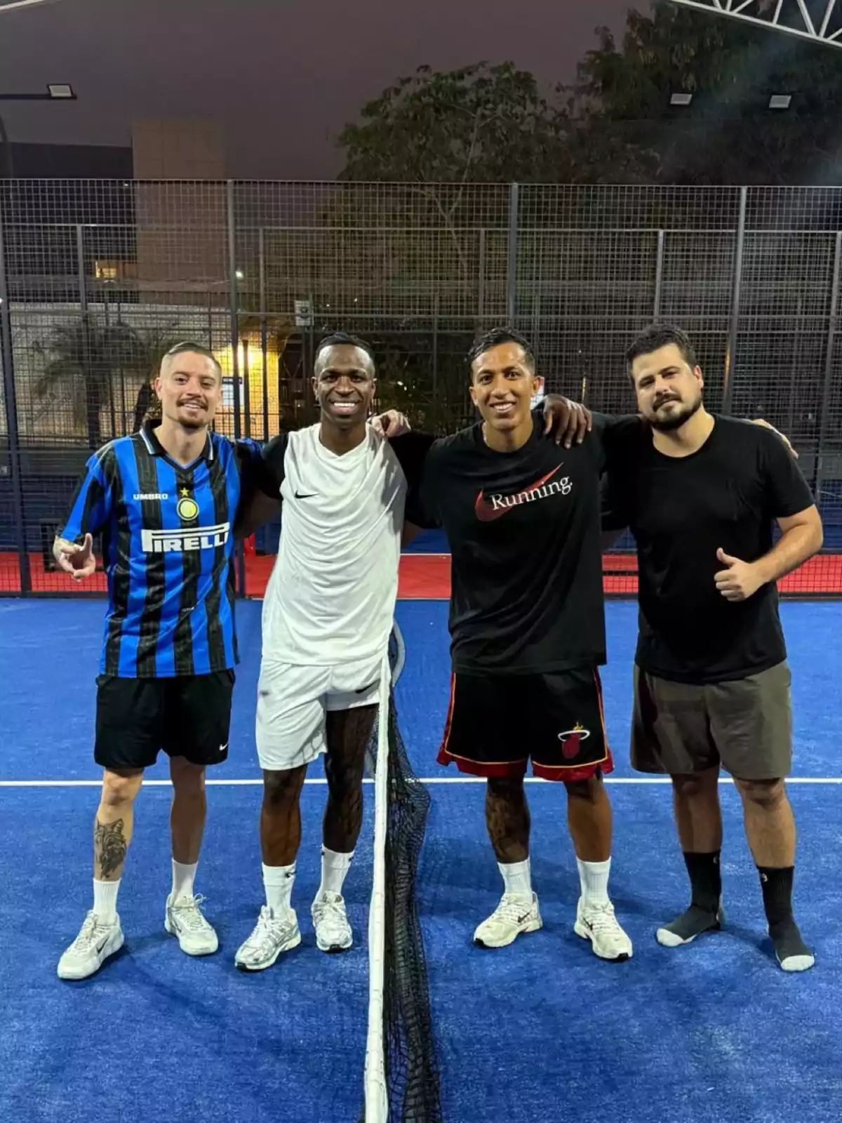 Cuatro hombres posan sonrientes en una cancha de pádel con una red en el centro y fondo de malla metálica