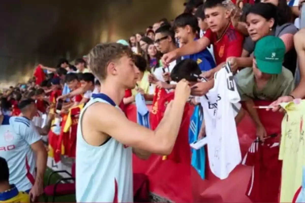 Un jugador de fútbol firma autógrafos a varios aficionados que le entregan camisetas y otros objetos desde las gradas.