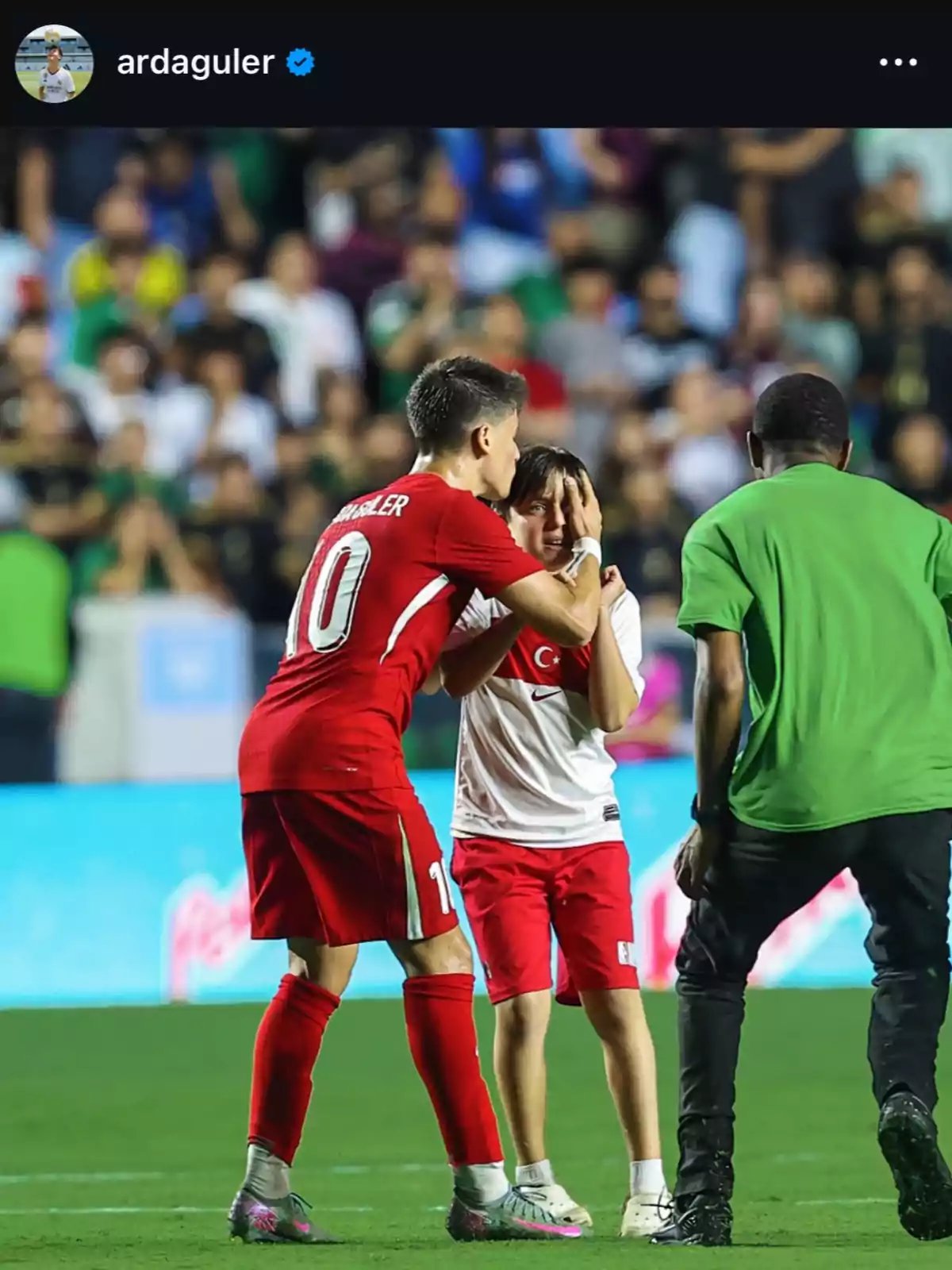 Jugador de fútbol con uniforme rojo abraza y consuela a un niño emocionado en el campo mientras un guardia se acerca Jugador de fútbol con uniforme rojo abraza y consuela a un niño emocionado en el campo mientras un guardia se acerca