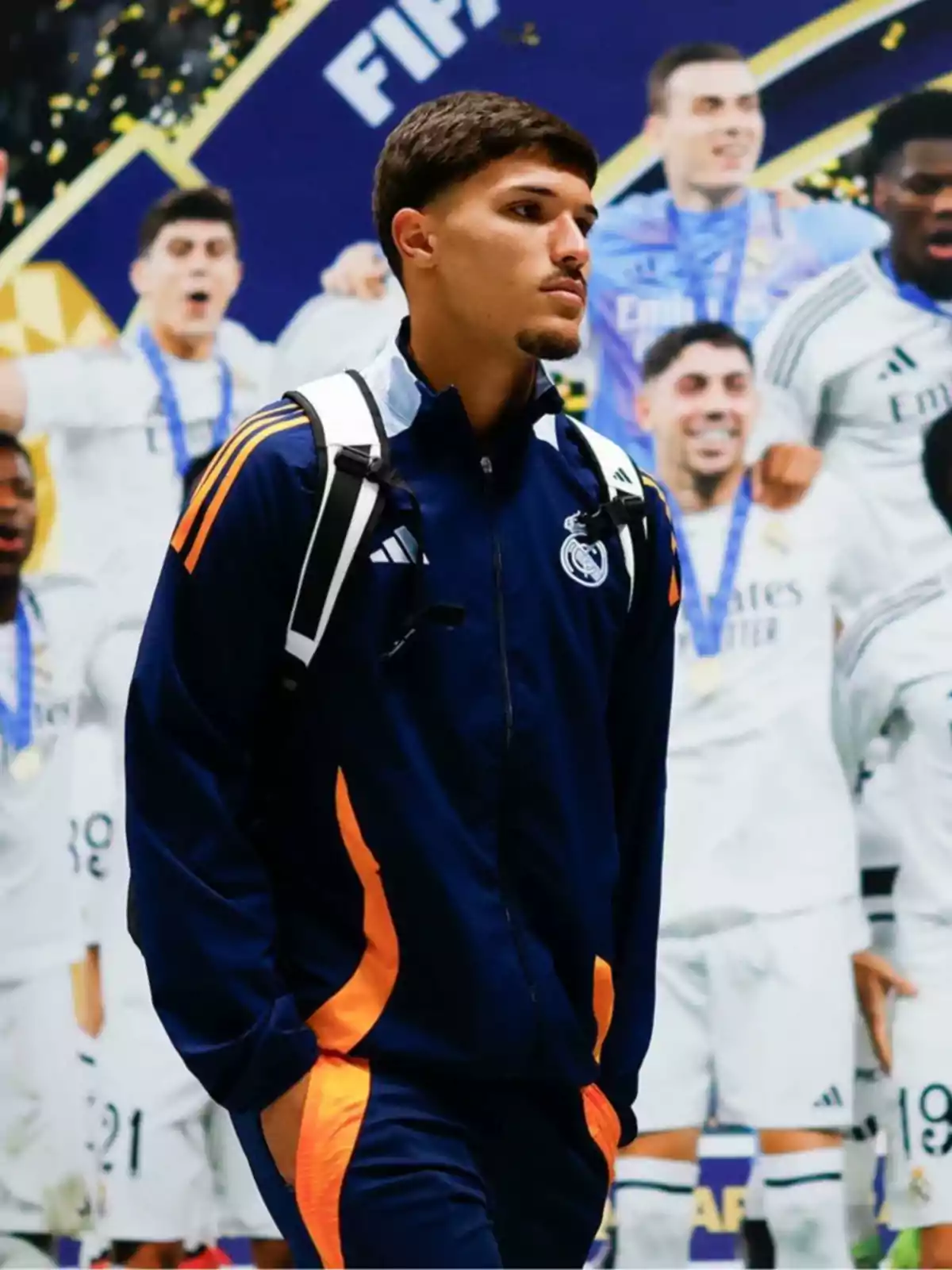 Joven con chándal azul marino y detalles naranjas del Real Madrid caminando con una mochila al hombro frente a un mural de jugadores celebrando Joven con chándal azul marino y detalles naranjas del Real Madrid caminando con una mochila al hombro frente a un mural de jugadores celebrando