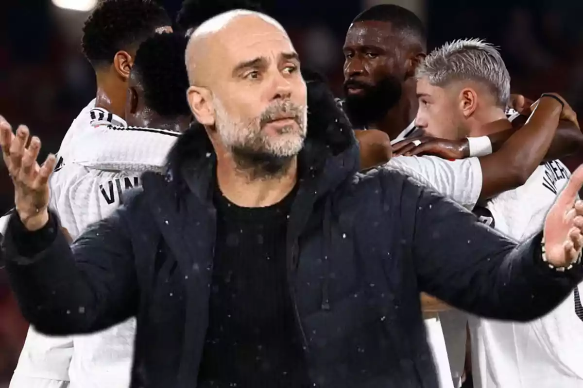 A man in a dark jacket gestures while a group of soccer players hug in the background.