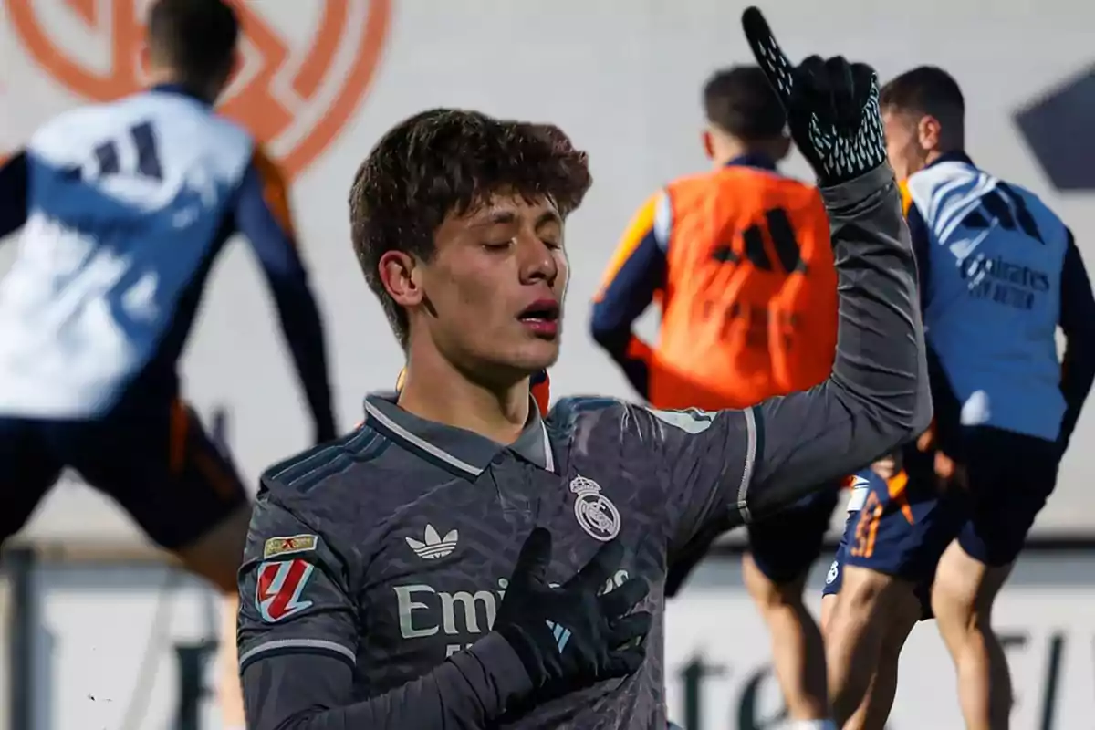 A soccer player in a grey uniform celebrates with his hand on his chest and his finger pointing up while other players train in the background.