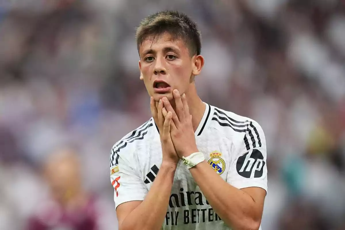A player with an expression of surprise or concern on the field, wearing a white uniform with black details and a team crest on the chest. A player with an expression of surprise or concern on the field, wearing a white uniform with black details and a team crest on the chest.
