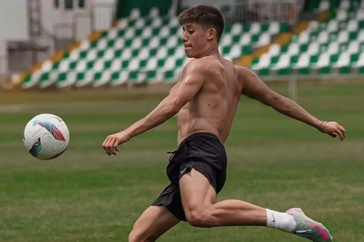 Joven sin camiseta jugando fútbol en una cancha de pasto mientras se prepara para patear el balón Joven sin camiseta jugando fútbol en una cancha de pasto mientras se prepara para patear el balón