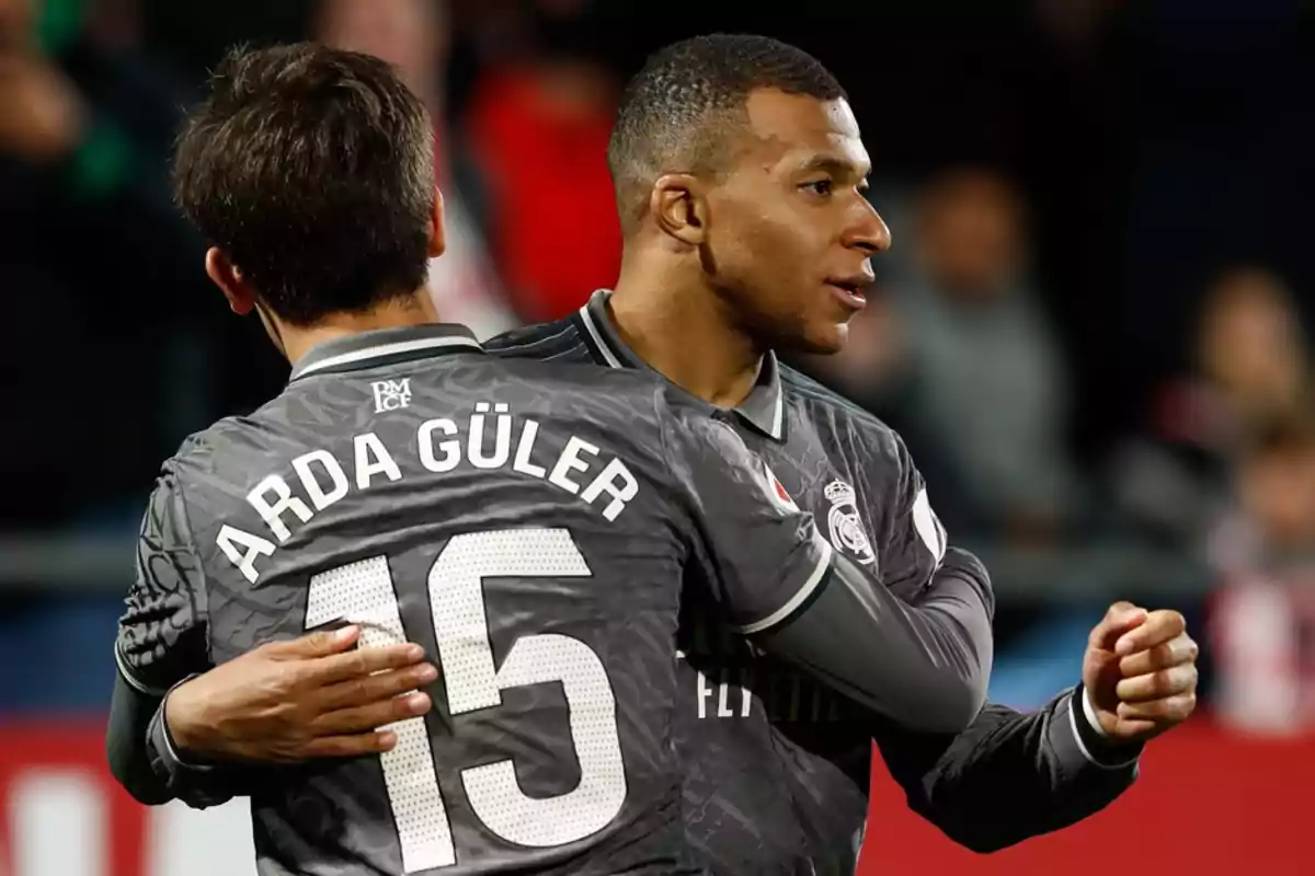 Two soccer players in gray uniforms embrace on the field.