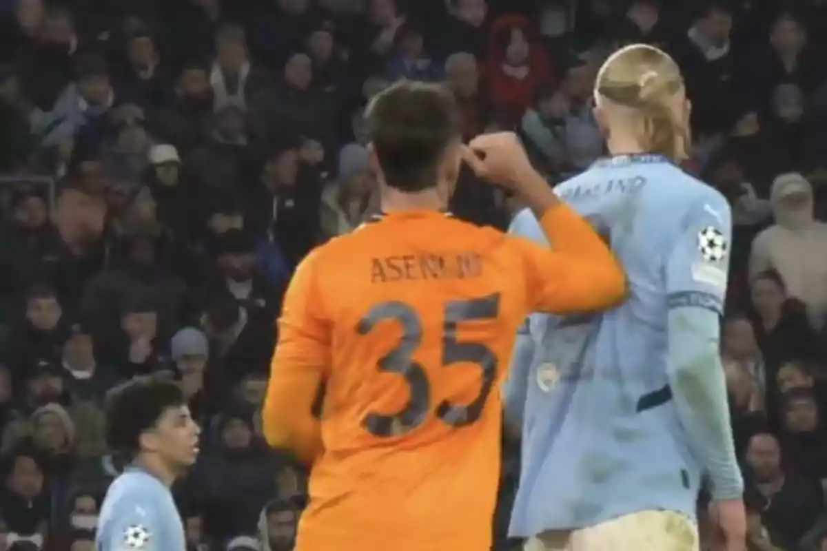 A player wearing an orange jersey with the number 35 is on the field next to another player in a light blue jersey, surrounded by spectators in the stands.