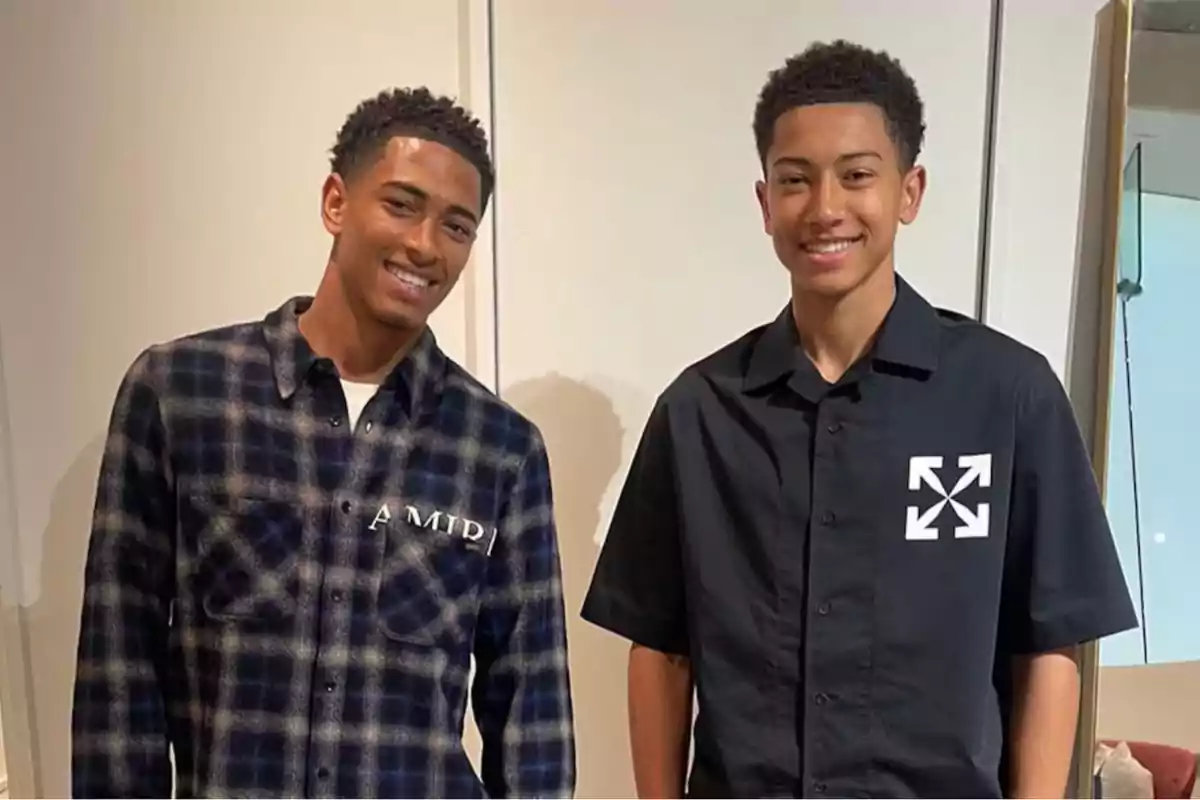 Dos jóvenes sonrientes posan juntos frente a una pared blanca, uno lleva una camisa de cuadros y el otro una camisa negra con un diseño gráfico.