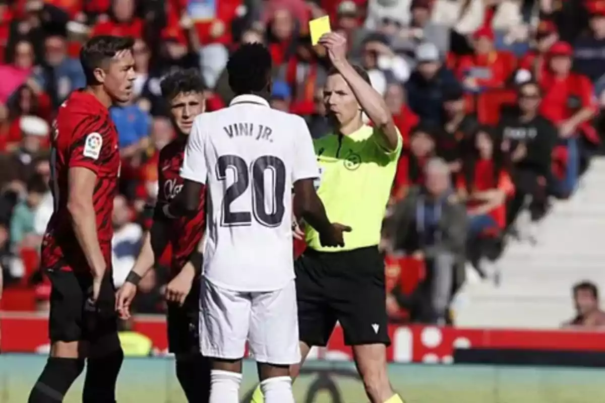 A referee shows a yellow card to a player with the number 20 on his white jersey while other players watch on the field. A referee shows a yellow card to a player with the number 20 on his white jersey while other players watch on the field.