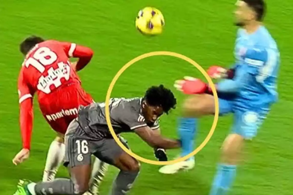 A soccer player in a gray uniform is accidentally hit by the foot of a goalkeeper dressed in blue while another player in a red uniform stands nearby.