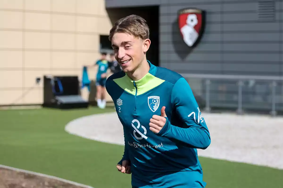 A smiling player in a blue and green training uniform runs outdoors near a building with a team logo on the wall. A smiling player in a blue and green training uniform runs outdoors near a building with a team logo on the wall.