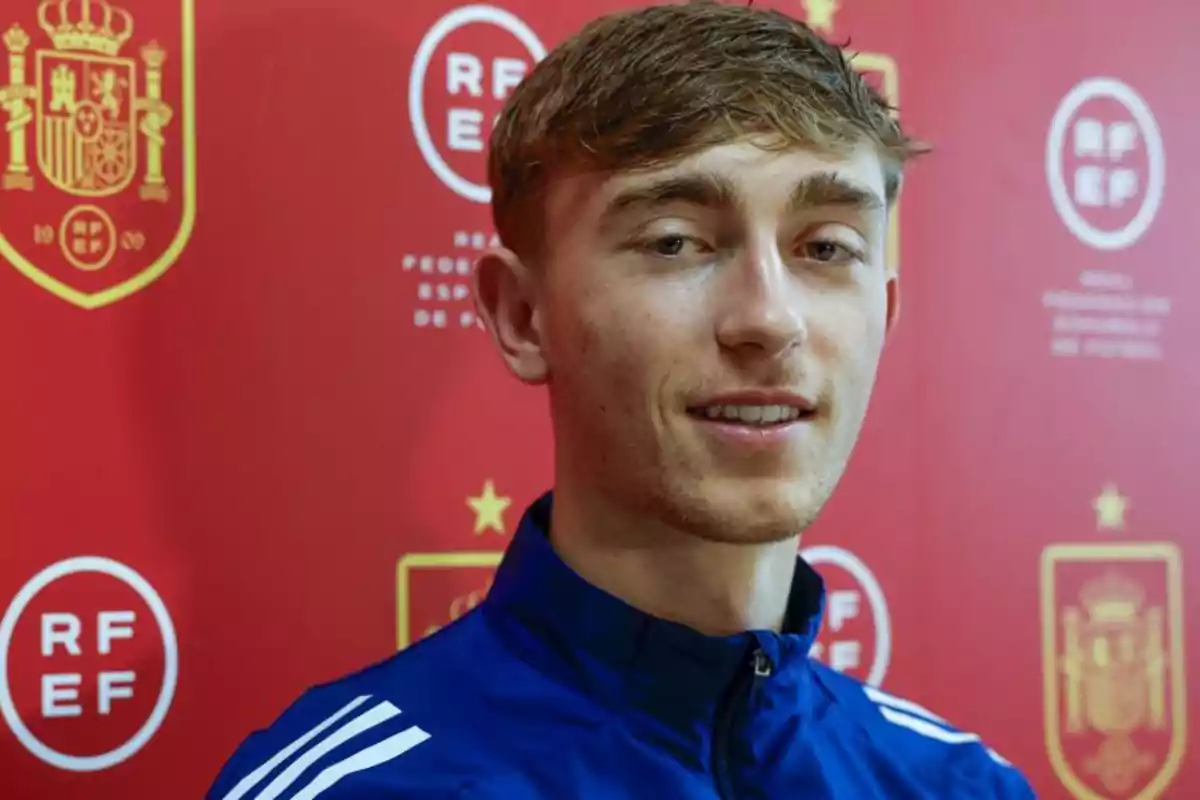 A young man in a blue jacket in front of a red background with logos of the Royal Spanish Football Federation. A young man in a blue jacket in front of a red background with logos of the Royal Spanish Football Federation.