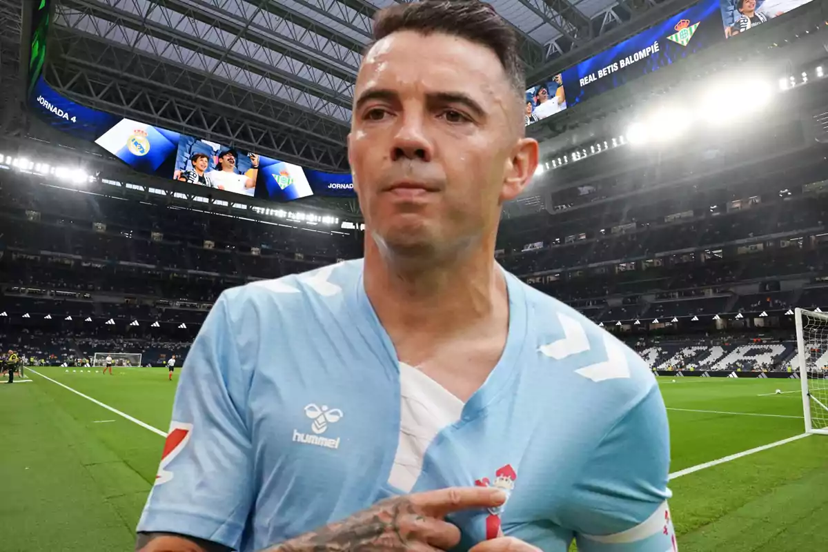 Un jugador de fútbol con una camiseta celeste está en un estadio con gradas y pantallas grandes al fondo.