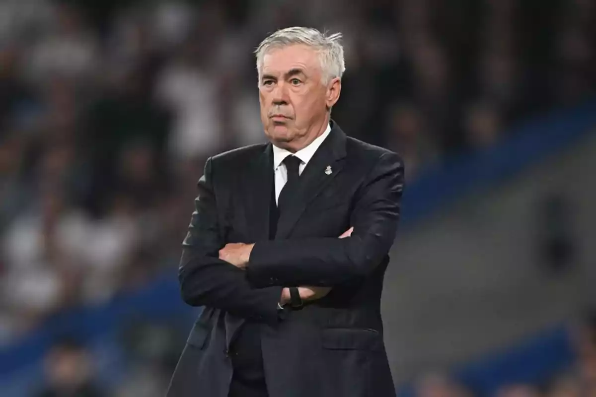 Un hombre de cabello canoso y traje oscuro con los brazos cruzados en un entorno deportivo. Un hombre de cabello canoso y traje oscuro con los brazos cruzados en un entorno deportivo.
