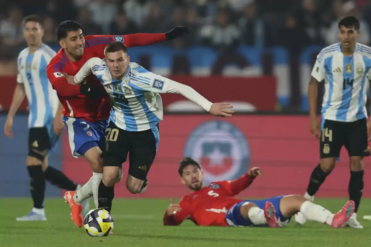 An Argentine player moves forward with the ball while being held by a Chilean opponent during a soccer match, with other players watching the play in the background. An Argentine player moves forward with the ball while being held by a Chilean opponent during a soccer match, with other players watching the play in the background.