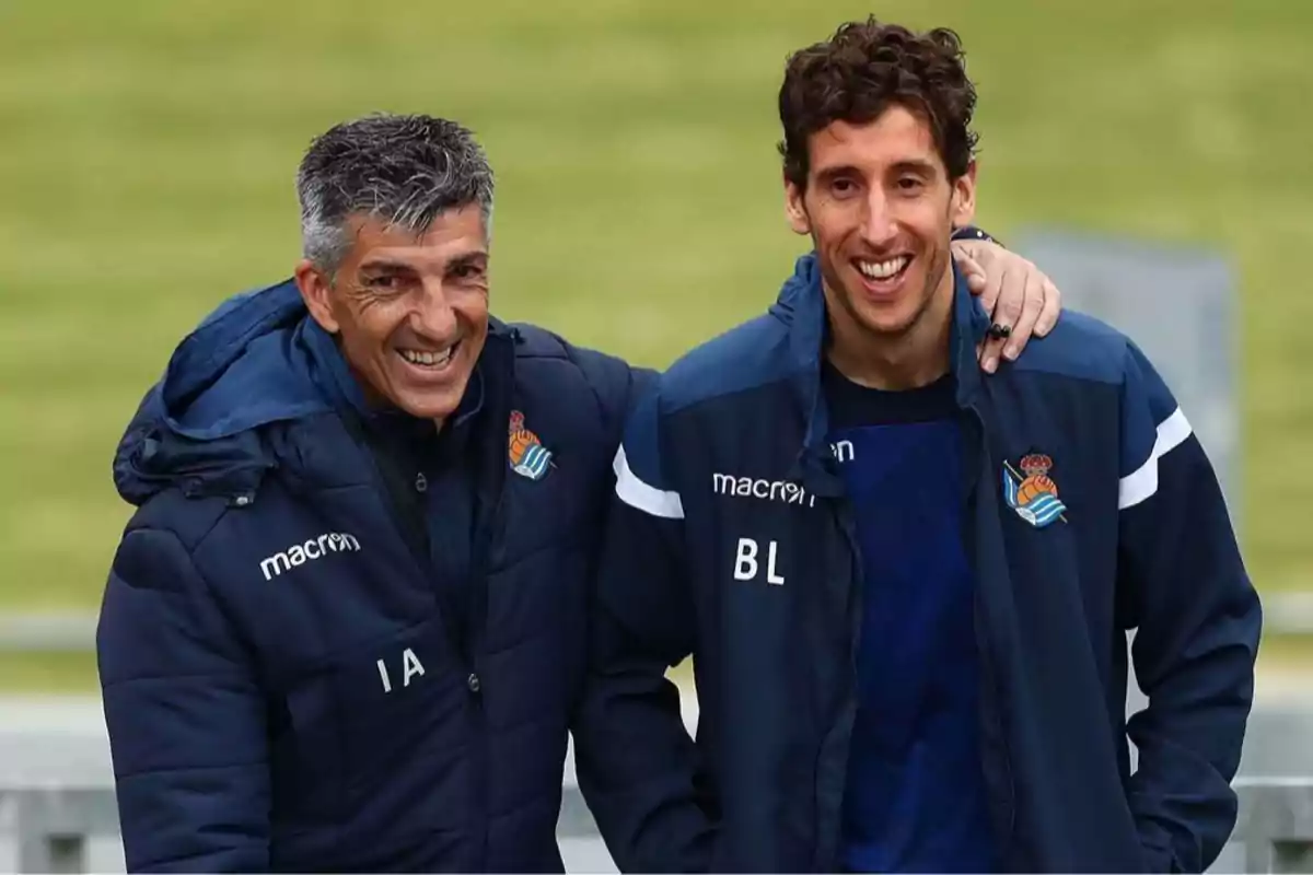 Two men smile while standing together, wearing sports jackets with a soccer team’s logos. Two men smile while standing together, wearing sports jackets with a soccer team’s logos.