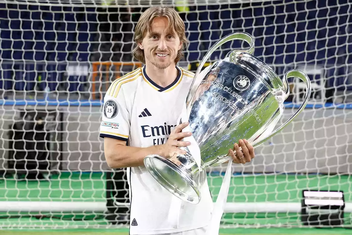 Un jugador de fútbol con uniforme blanco sostiene un gran trofeo plateado frente a una portería.