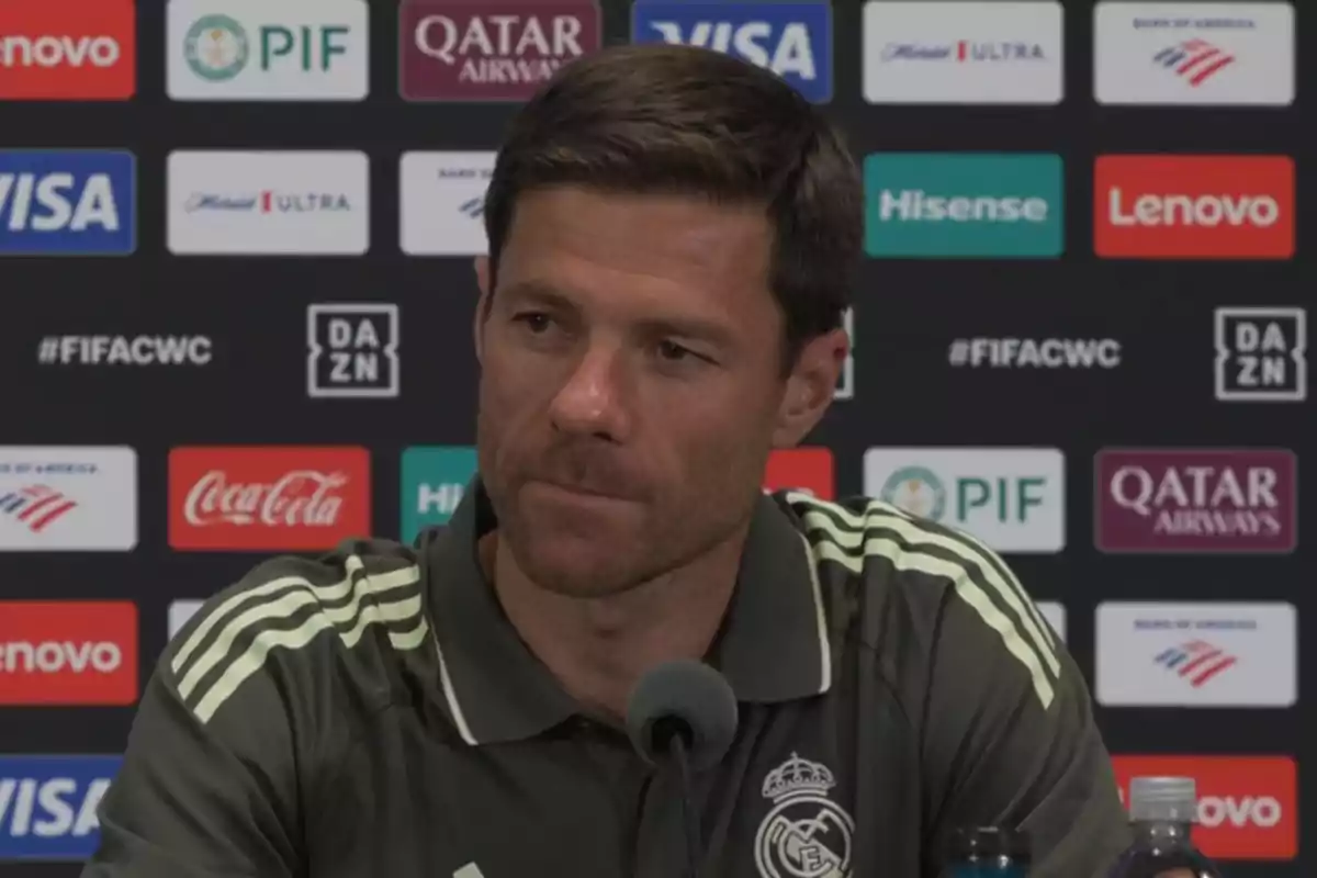 Man wearing a Real Madrid training jersey sitting in front of a microphone at a press conference with a sponsor backdrop Man wearing a Real Madrid training jersey sitting in front of a microphone at a press conference with a sponsor backdrop