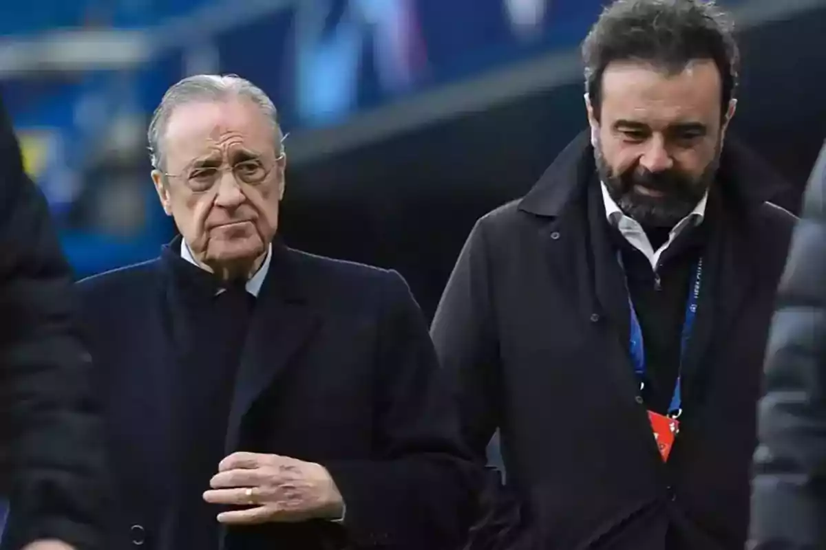 Two men walking together in an outdoor setting, both wearing dark coats and with serious expressions. Two men walking together in an outdoor setting, both wearing dark coats and with serious expressions.