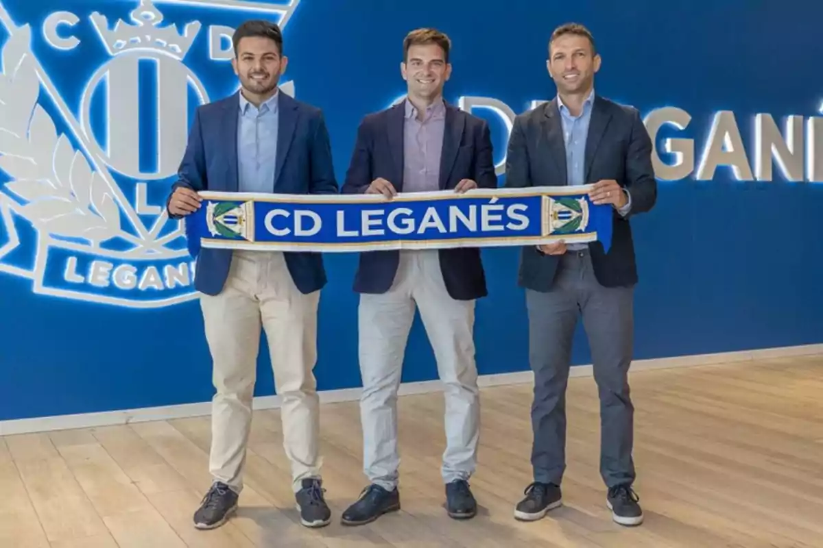 Three men stand smiling, holding a blue CD Leganés scarf in front of a blue wall with the club’s crest and name. Three men stand smiling, holding a blue CD Leganés scarf in front of a blue wall with the club’s crest and name.