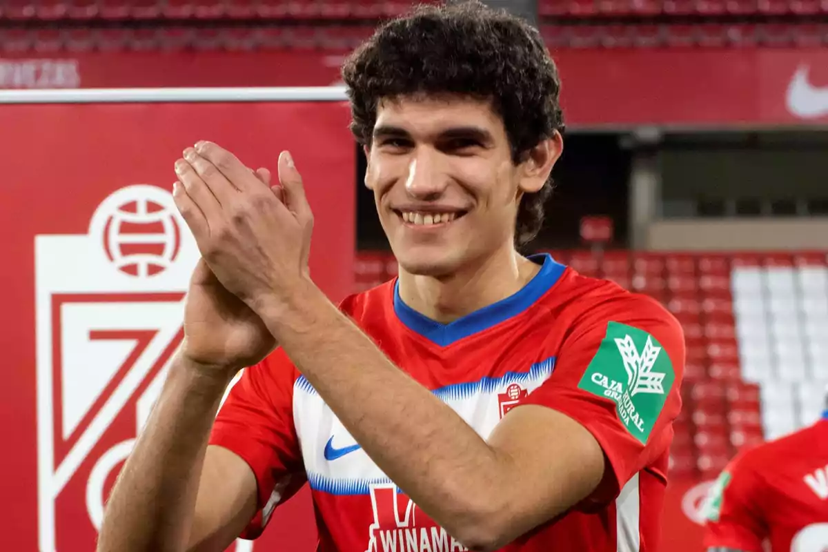 A soccer player applauding in a stadium wearing a red jersey.