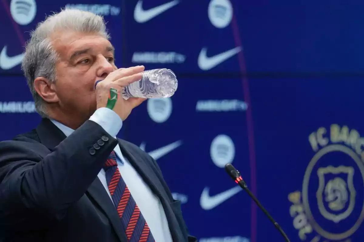 A man in a suit drinks water from a bottle during a press conference with logos in the background. A man in a suit drinks water from a bottle during a press conference with logos in the background.