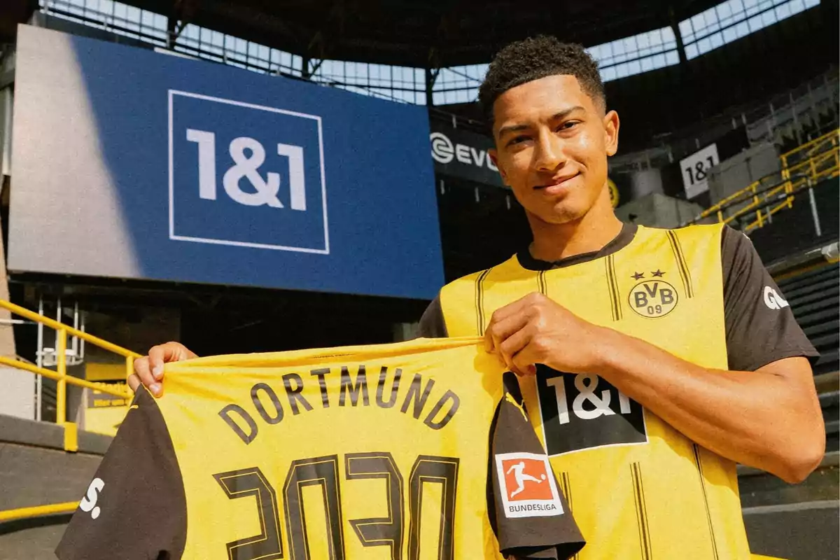Jugador del Borussia Dortmund sonríe mientras sostiene una camiseta amarilla con el nombre del equipo y el número 2030 en un estadio de fútbol Jugador del Borussia Dortmund sonríe mientras sostiene una camiseta amarilla con el nombre del equipo y el número 2030 en un estadio de fútbol
