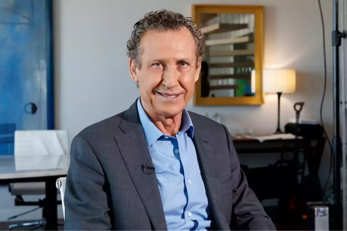 A man with curly, gray hair smiles as he sits in an office with a blue painting and a mirror on the wall. A man with curly, gray hair smiles as he sits in an office with a blue painting and a mirror on the wall.