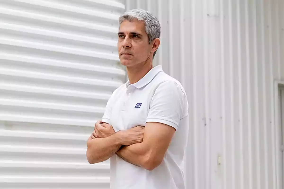 Gray-haired man wearing a white polo jersey standing in front of a white metal wall with his arms crossed Gray-haired man wearing a white polo jersey standing in front of a white metal wall with his arms crossed