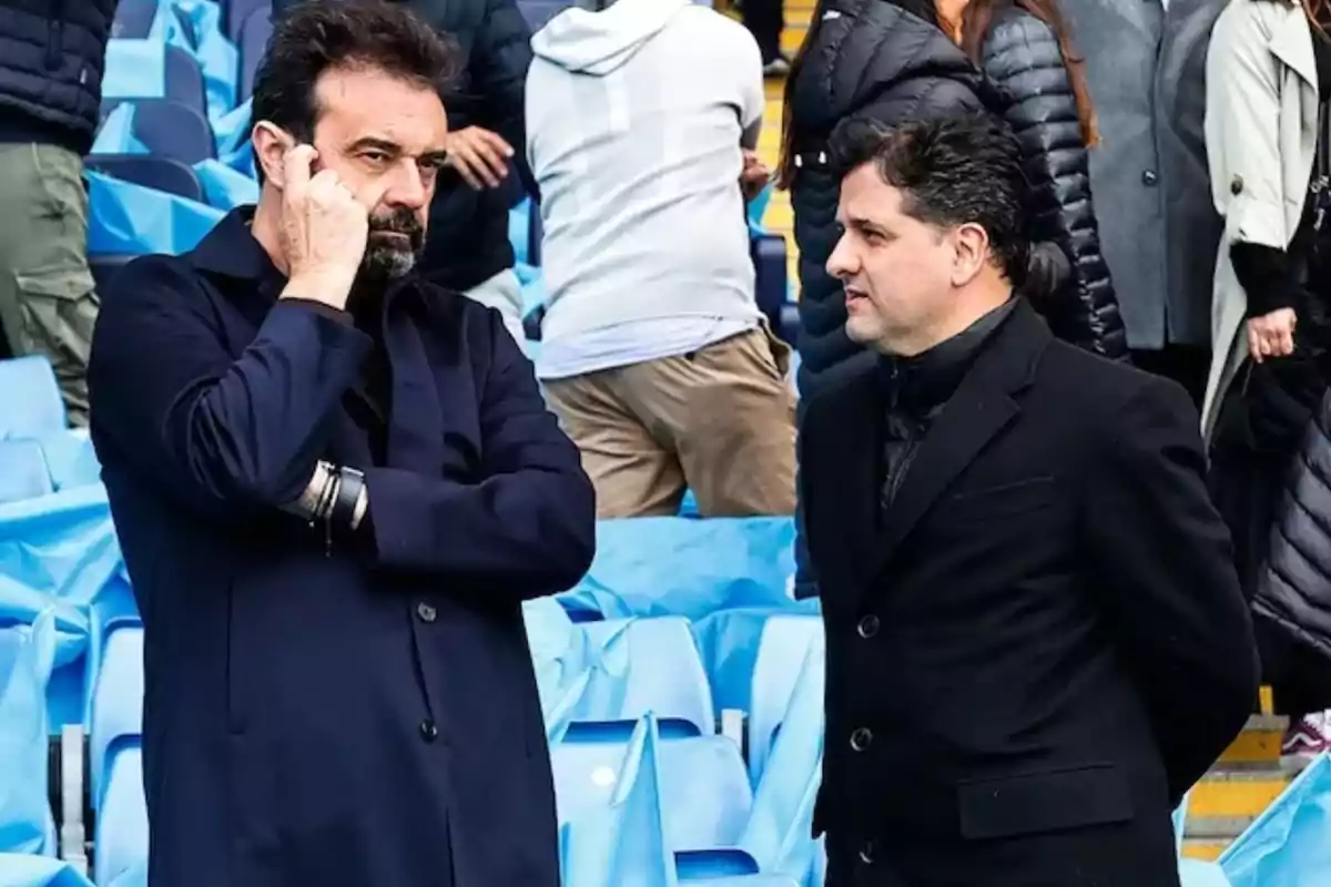Two men standing in a stadium, one talking on the phone and the other listening intently, surrounded by empty seats and people in the background. Two men standing in a stadium, one talking on the phone and the other listening intently, surrounded by empty seats and people in the background.