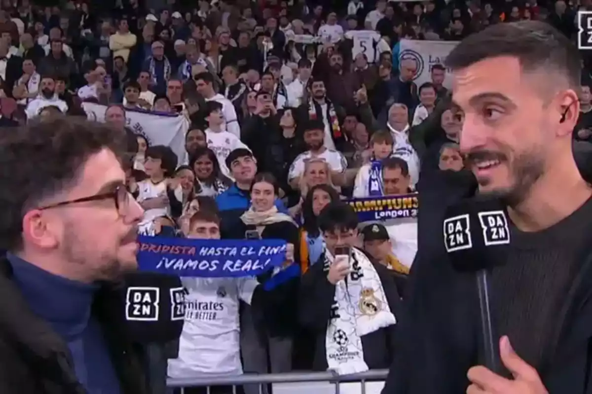 Dos personas con micrófonos de DAZN están hablando frente a una multitud de aficionados que sostienen bufandas y pancartas en un evento deportivo.