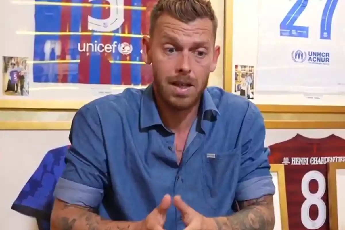 A man in a blue jersey sits in front of framed soccer jerseys on the wall. A man in a blue jersey sits in front of framed soccer jerseys on the wall.