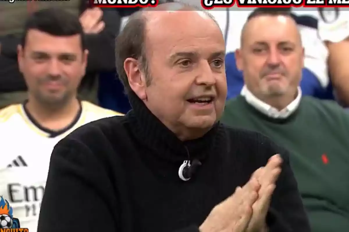 A man in a black sweater is speaking on a TV show with people sitting behind him.