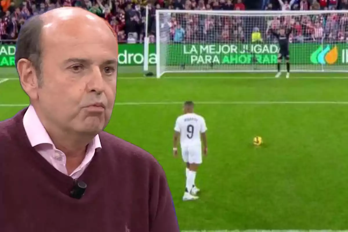 A man in a burgundy sweater appears in the foreground while in the background a footballer can be seen preparing to take a penalty in a stadium full of spectators.