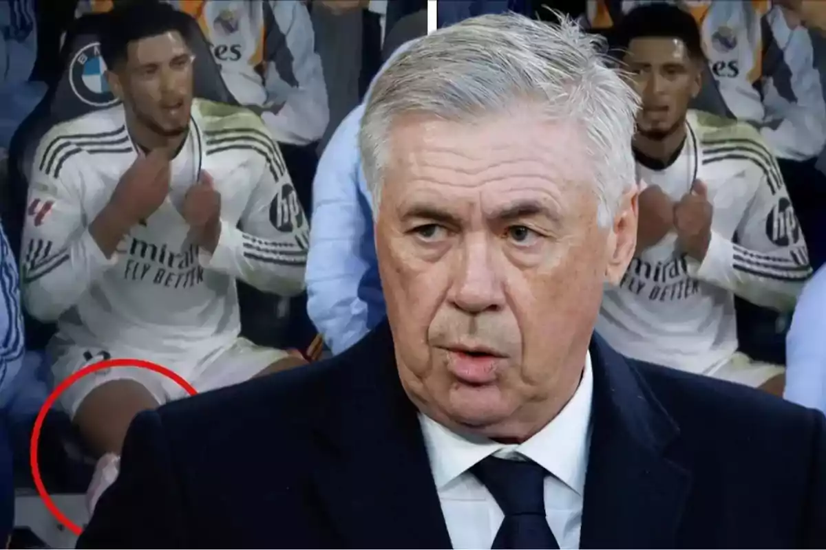 A gray-haired man in the foreground with a serious expression and a player in the background sitting on the bench in a white uniform.